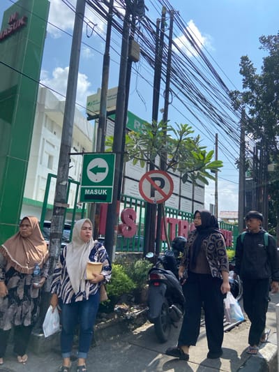 The image shows a street scene in Cimahi, Indonesia (latitude: -6.884, longitude: 107.553).  Three women and one man are standing on the sidewalk in front of a building. The women are wearing traditional Muslim clothing, and the man is wearing a black jacket and jeans. Two of the women are holding shopping bags, and the other is holding a book or folder. A motorcycle is parked on the sidewalk, and several power lines are visible overhead.  A green sign with an arrow pointing left reads "MASUK", which translates to "enter". A red sign with the letter "P" circled in white is visible behind the motorcycle. The sign prohibits parking.  The building behind the people is a large, modern structure with a green facade.  The building is named "RS." There are also green plants and trees on the sidewalk and in front of the building.  The sky is clear and blue with some clouds.  The scene is one of a normal urban street scene in a developing country.