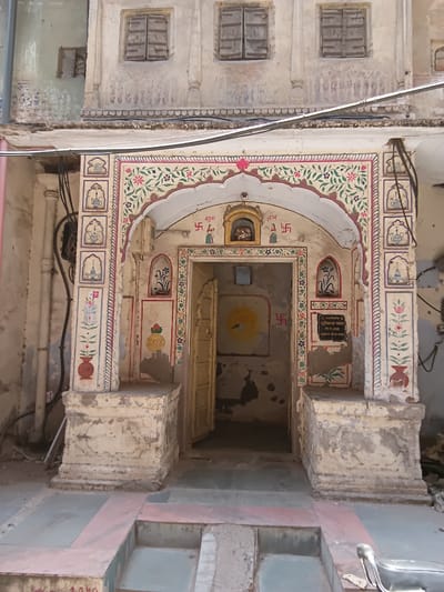 Here's a detailed description of the image:

The image captures a doorway in what appears to be the city of Jaipur, India, known for its historic architecture. The scene is vertically oriented.

**Subject:** The focal point is an ornate doorway leading into an interior space.

**Objects and Details:**

*   **Doorway:** The doorway is embellished with traditional Indian floral and geometric designs in vibrant colors (red, green, pink) on a light background. The paint shows signs of aging and weathering. Swastikas appear near the top corners, next to the words "Shubh" and "Labh"

*   **Inscription:** Above the doorway is a small rectangular inscription plaque, written in Hindi and including the word "Bhawan".

*   **Building Exterior:** The adjacent exterior wall has a series of small, shuttered windows. The walls are painted in a pale, neutral color and show wear.

*   **Wires:** A bundle of electrical wires runs across the top of the doorway.

*   **Interior:** A glimpse into the interior reveals a poorly lit space with an open door to a third room.

*   **Floor:** The foreground features a floor made of stone or tile slabs in shades of gray, pink, and green.

**Environment:** The environment seems to be an older part of the city, given the aged condition of the building. The lighting suggests an outdoor shot, though the doorway could be partially sheltered.