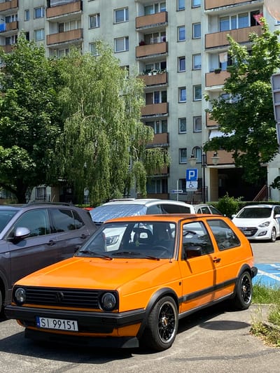 The image shows an orange Volkswagen Golf Mk2 parked on an asphalt surface, likely a parking lot. The car is positioned in the foreground, taking up a significant portion of the frame. It has black trim around the wheel arches and black rims with silver accents. The license plate is visible and reads "SI 99151".
In the background, a multi-story residential building with light-colored walls and balconies is visible. Green trees with dense foliage are present between the building and the parking area, partially obscuring the lower floors of the building. To the right of the orange car, a white Hyundai i30 is parked.
Signs are visible on a lamppost, including a blue parking sign with a white "P" symbol and a wheelchair accessibility symbol. Another sign below it appears to be a street name or directional sign with Polish text.
The scene appears to be an urban environment, likely a residential area in Bielsko-Biała, Poland, given the car's license plate format and the architectural style of the building. The lighting suggests it is daytime with bright sunlight casting shadows. No people are actively visible in the scene.