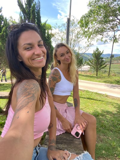 Two women are posing for a selfie outdoors. The woman in the foreground on the left has dark hair and is wearing a pink tank top. She has tattoos on her arms and neck. The woman on the right has blonde hair and is wearing a white crop top and a pink pleated skirt. She is holding a pink phone case. They are sitting on a wooden bench in a grassy area with trees and hills in the background. The location is identified as Thôn 1, Vietnam.