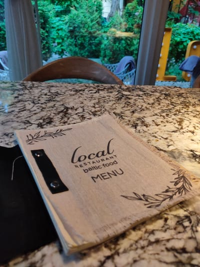 A menu is on a granite countertop in a restaurant with an outdoor seating area visible through a window. The menu is light brown with decorative green leaves printed on it. The text on the menu reads "local RESTAURANT baltic food MENU". A dark object, possibly a placemat or napkin, is tucked under the menu. In the background, through the window, lush green trees and outdoor seating with blue cushions are visible. Large illuminated letters spelling "LOVE" are also visible in the distance, positioned to the right. The overall ambiance suggests a casual dining experience in a garden setting, likely in Svetlogorsk, Russia, a resort town known for its coastal location and German architectural influences, which could explain the prominent "LOVE" sign.