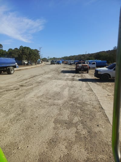 The image shows a dusty, unpaved road in El Valle del Espíritu Santo, Venezuela. Several vehicles are present, including a large blue tanker truck on the left, a red and white pickup truck, a white pickup truck, and other vehicles further in the background. Some of these vehicles appear to be water trucks, indicated by the tanks on their backs. The road is dry and uneven, with scattered debris. The environment is arid, with sparse vegetation and hills in the background. The sky is clear and blue with a few wispy clouds. There are power lines visible overhead. The scene appears to be in a somewhat industrial or logistical area, possibly related to water distribution.