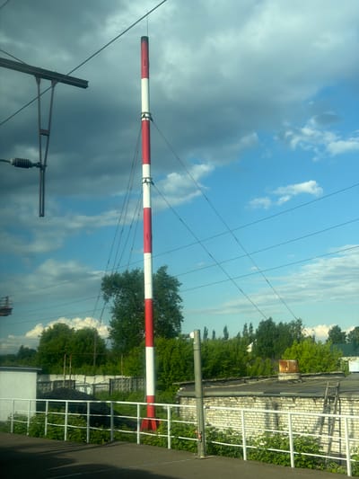 In Vyatskiye Polyany, Russia, a red and white striped pole stands tall against a backdrop of blue sky with scattered clouds. The pole is a prominent vertical element in the image. In the foreground, a white railing separates a paved area from a strip of green vegetation. Behind the railing, there is a white building with a dark roof and a section of a white brick wall. Further back, more buildings with dark roofs are visible, along with a pile of dark material, possibly coal, and a metal fence. Trees and foliage are scattered throughout the scene, suggesting a semi-urban or industrial setting. Overhead, power lines crisscross the sky, and to the left, a portion of an overhead railway structure is visible. The overall atmosphere is bright and appears to be daytime.