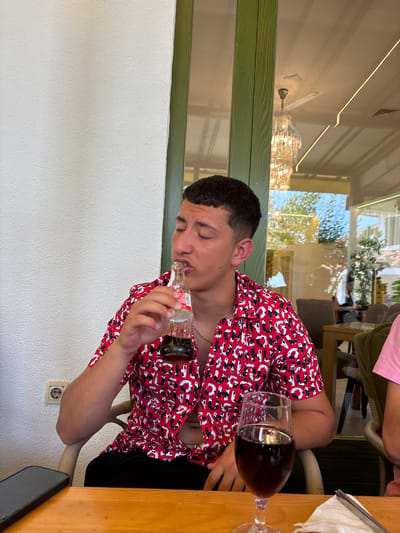A young man in a red and white printed shirt is drinking directly from a glass Coca-Cola bottle at an outdoor cafe. A glass of dark liquid, possibly Coca-Cola or another beverage, sits on the wooden table to his right. The location appears to be an open-air restaurant or cafe with large windows reflecting a bright outdoor environment, featuring trees and possibly other buildings. The man is seated at a table with other patrons nearby. Based on the user's prompt, this is likely located in Velika Plana Municipality, Serbia.