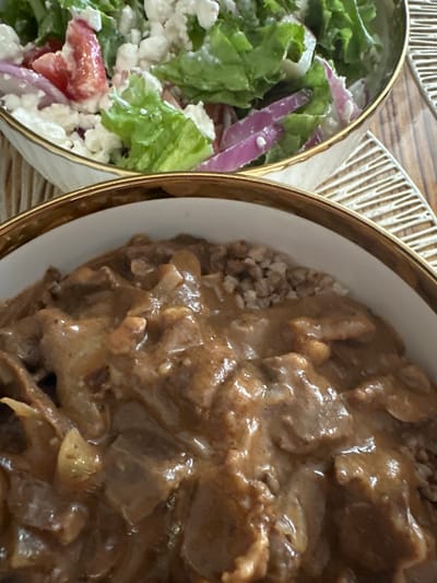 The image shows a close-up view of two bowls of food, likely part of a meal. In the left bowl, there is a salad consisting of lettuce, red onion, tomatoes, and crumbled white cheese, possibly feta. The salad appears to be dressed with a creamy white dressing. The bowl itself has a golden rim. To the right, a second bowl contains a dish of meat in a rich, brown sauce, served over a bed of what appears to be buckwheat groats. The meat is cut into bite-sized pieces and is coated in the thick sauce. This bowl also has a matching golden rim. Both bowls are placed on a wooden surface, with a textured placemat visible in the background. The lighting suggests it is daytime. There is no visible text in the image.