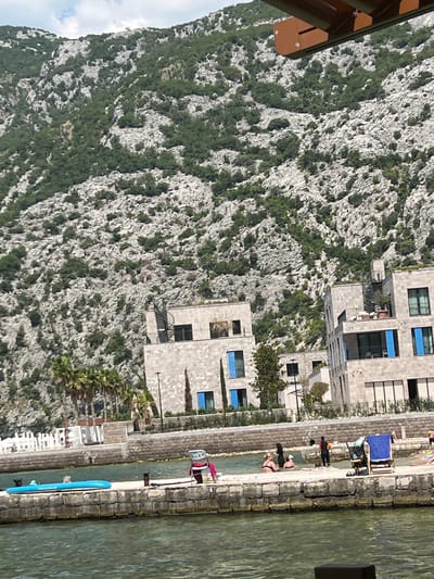 The image depicts a waterside scene with a steep, rocky mountainside covered in sparse green vegetation dominating the left side. On the right, a modern building with several floors and prominent blue-tinted windows is situated along a stone-paved waterfront. People are visible on the waterfront, some lounging or sunbathing, while others appear to be engaged in activities near the water. A blue paddleboard rests on the edge of the water. The water itself is calm and greenish, with sunlight reflecting off its surface. In the foreground, a section of a brown wooden structure is visible, likely part of a balcony or overhead covering. The scene suggests a sunny day, possibly in the afternoon, given the lighting and the presence of people enjoying outdoor leisure activities. The overall setting appears to be a resort or residential area along a body of water, with the dramatic mountain backdrop providing a striking contrast to the man-made structures. No visible text is present in the image.