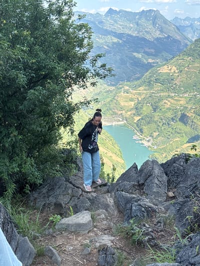 A woman sits on the rocky edge of a cliff. She is wearing a dark t-shirt, light blue jeans, and sandals. She appears to be looking at something in her hand. Lush green trees and vegetation are in the foreground, partly obscuring the view. A vast mountainous landscape stretches out behind her, with terraced fields and a body of water visible. This landscape indicates the location is likely within the Đồng Văn Commune, Vietnam. The cliffs appear rocky and naturally formed, indicative of a mountainous environment.