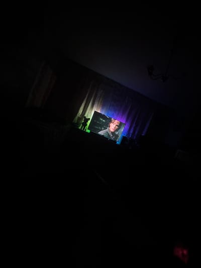 A dark, low-angle shot captures a dimly lit room with a television screen displaying a man's face. The TV is positioned on a surface, and behind it, curtains with a vertical pattern are visible. In front of the TV, a small, dark figure, possibly a figurine, stands. The room is otherwise enveloped in shadows, with only a faint glow from the television and what appears to be ambient lighting behind the curtains, casting a gradient of colors from blue to purple. A chandelier is silhouetted against the ceiling in the upper right corner of the frame. The image appears to be taken in a residential setting, likely a living room, within the city of Makiivka, Ukraine.