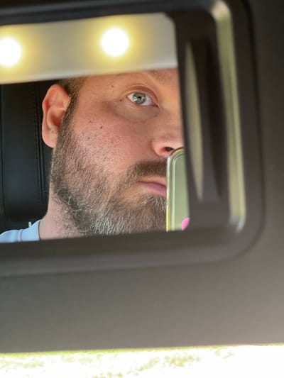 A close-up, eye-level, medium shot is captured in the reflection of a vehicle's visor mirror. The mirror is lit from above by two bright circular lights. A Caucasian male with blue eyes and a beard is reflected in the mirror. His left eye and the left side of his face are visible, along with his ear. A portion of a mobile phone, with a reflective screen and what appears to be a pink case, is visible in the right side of the mirror, held by the man. The man is wearing a light-colored t-shirt. The background of the reflection shows the interior of a vehicle, with a black seat visible behind the man. The visor frame, in a dark grey or black, surrounds the reflection. Outside the vehicle, bright sunlight illuminates a blurred green area, suggesting an outdoor environment, possibly grass or foliage. The location is not specified in the image, and there is no indication that it is in Старый Кыч, Russia.