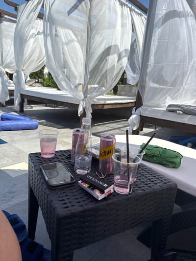 The image shows a collection of used drinks and a mobile phone on a black rattan table next to a white canopy bed. Two clear plastic cups with straws and remnants of pink liquid are visible, along with an opened can of Schweppes Pink Tonic & Elderflower. A partially empty bottle of water with a label in Cyrillic script is also on the table. A pack of cigarettes with visible text and a phone with a reflective screen are also present. In the background, a green folded cloth rests on a white surface, and part of a wooden structure supporting the canopy is visible. The overall scene suggests a relaxed setting, possibly outdoors by a pool or at a resort, with the time of day appearing to be daytime with bright sunlight casting shadows. The location is Tsigov Chark, Bulgaria.