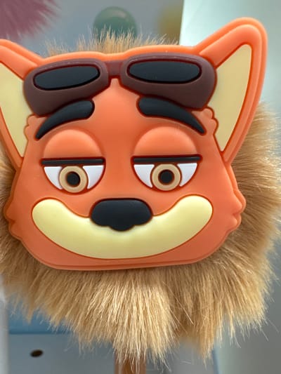 This is a close-up, vertically oriented photograph of an orange, cartoonish animal face with a fluffy brown mane. The animal appears to be a fox or a lion cub, with large, exaggerated features. It wears oversized, dark brown sunglasses with black lenses, which obscure its eyes partially. Two prominent, black, curved shapes suggest eyebrows or perhaps part of the sunglasses' design. Below the sunglasses, the face has two round eyes, each with a white sclera, a light brown iris, and a black pupil. The face is framed by a pale yellow outline, and the orange plastic material forms the main facial features. The right side of the image is dominated by a dense, light brown, furry mane, giving the impression of a plush toy or a character with a distinctive hairstyle. The background is blurred and consists of soft, muted colors, hinting at an indoor setting, possibly a desk or shelf, with a light blue element visible on the right. The overall lighting is bright, suggesting daytime or well-lit indoor conditions. No text is visible in the image. The image composition focuses solely on the character's face and mane, creating a playful and whimsical impression.