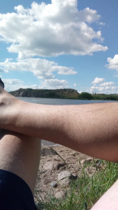 The media file is a close-up shot of a person's arms and legs, positioned in the foreground against a scenic backdrop of a lake and a hilly landscape. The person's skin is visible, with fine hairs on the forearm. They are wearing dark blue shorts or pants. The setting appears to be a natural environment near water. The ground in the foreground is covered with small stones, sand, and patches of green grass, suggesting a lakeside or riverbank location.

In the background, a calm body of water stretches across the frame, with a prominent hill or small mountain covered in dark green vegetation rising from the opposite shore. The sky above is a bright blue, dotted with fluffy white clouds, indicating fair weather. The overall impression is one of a relaxed outdoor scene, possibly during daytime due to the bright lighting and blue sky. There are no visible text elements in the media file.