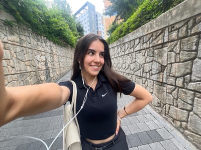 A young woman with long, dark hair is taking a selfie outdoors. She is wearing a black crop top with a Nike logo and black pants. She has white earbuds in her ears and is holding a light-colored purse over her shoulder. Her left arm is raised to take the photo, and her right hand is resting on her stomach. She is smiling and looking at the camera.
The background shows a cobblestone pathway and a stone wall. There are also some lush green trees and a modern, multi-story building with Korean text visible on its side. The lighting suggests it is daytime, likely a bright day given the shadows. The overall scene appears to be an urban or semi-urban environment, possibly a university campus given the building and well-maintained pathway. The woman's expression is cheerful and relaxed.