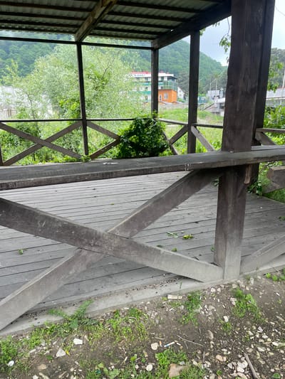 The image depicts a partially covered wooden structure, likely a gazebo, in Olginka, Russia. The gazebo has a wooden deck and a cross-patterned railing. Beyond the railing, there's greenery, including trees and grass. In the distance, buildings are visible, including one with an orange exterior. The ground outside the gazebo is covered with dirt, small rocks, and some scattered debris. The photograph appears to have been taken from inside the gazebo, looking outward.