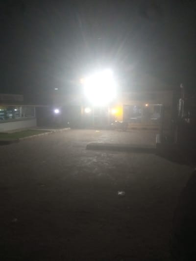 The image shows a poorly lit outdoor scene at night, likely in a commercial area. A bright, overexposed light source dominates the upper center of the frame, making it difficult to discern details. To the left, a building with windows is partially visible, suggesting a shop or office. A paved area, possibly a parking lot or walkway, is in the foreground, with some wet patches indicating recent rain or moisture. A motorcycle is parked in the middle ground, bathed in the light. The overall atmosphere is dark and somewhat hazy, with limited visibility. The location is identified as Television, Nigeria, but no specific landmarks are discernible from the image itself.