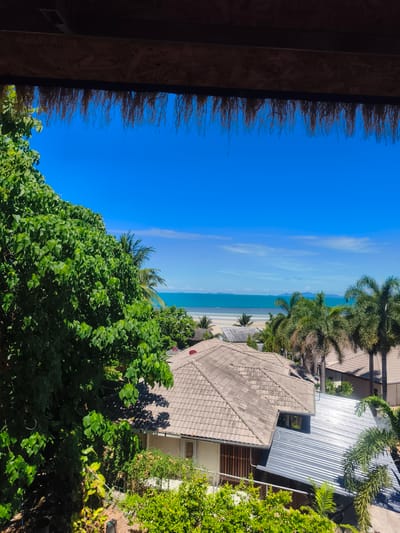 The image depicts a tropical scene, likely in Baan Thong Po, Thailand, with a clear blue sky dominating the upper portion. In the foreground, a thatched roof overhang partially obscures the view, with dried palm fronds hanging down. Lush green foliage, primarily from a large tree on the left, fills the lower left of the frame.
Beyond the vegetation, a cluster of buildings with tiled roofs is visible. The tiles are predominantly a muted, earthy tone. One building has a distinctly modern, corrugated metal roof that gleams in the sunlight, contrasting with the older tiled roofs. Scattered palm trees with their characteristic fronds are interspersed among the buildings and along the coastline.
The middle ground features a sandy beach bordering a turquoise ocean. The water appears calm, with a gentle shoreline. In the distance, faint outlines of islands or landmasses can be seen against the horizon. The overall impression is one of a peaceful, sunny day in a coastal resort area. No people are present in the image, and no specific actions or interactions are depicted. The environment is characterized by vibrant natural elements and man-made structures characteristic of a tropical setting.