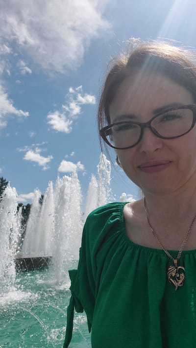 This is a selfie taken outdoors in front of a fountain on a sunny day. The subject is a woman wearing glasses and a green top, with a necklace featuring a heart-shaped pendant. She is smiling and looking towards the camera. In the background, multiple water jets from a fountain are visible, with trees and a blue sky with clouds also present. The scene suggests a public park or recreational area, possibly within the city of Komsomolsk-on-Amur, Russia.