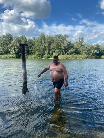 A man is wading into a body of water, likely a river or lake, near a tall, metal post sticking out of the water. He is shirtless and wearing dark shorts. The water is a deep blue and reflects the sky and surrounding greenery. Lush green trees line the distant bank of the water, and the sky above is partly cloudy with a mix of blue sky and white clouds. The overall setting appears to be natural and tranquil. The location is identified as Vodyane, Ukraine.