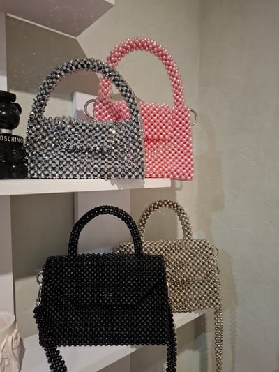This is a photograph of four beaded handbags displayed on white shelves. In the top row, two handbags are visible. The one on the left is silver and made of faceted beads, while the one on the right is pink and made of small, smooth pearls. Behind these, a black Moschino teddy bear perfume bottle is partially visible on the left. In the bottom row, two more handbags are displayed. The one on the left is black and made of smooth, round beads, and the one on the right is gold and also made of smooth, round beads. The background is a light-colored wall with a textured pattern. The lighting is soft, creating shadows on the wall. The image appears to have been taken indoors, likely in a retail or home setting. There are no people or animals in the image. The photo does not show any recognizable landmarks or context that would place it specifically in Korenovsk, Russia.