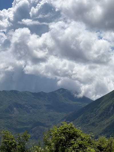 The image captures a vast, verdant mountainous landscape under a partly cloudy sky. The majority of the frame features towering mountains covered in dense, dark green foliage, indicative of a heavily forested region. The slopes appear steep and rugged.
Above the mountains, a dynamic sky is visible, filled with large, volumetric cumulus clouds. These clouds range from brilliant white, illuminated by sunlight, to darker grey patches, suggesting varying thickness and shadows. Patches of clear blue sky peek through the cloud cover.
In the extreme foreground, on the right side of the image, lush green foliage from a tree or bush partially frames the scene, hinting at the observer's position being within or very close to a forested area, looking out over the valley and opposing peaks.
The lighting suggests a bright daytime scene, likely late morning or early afternoon, with clear visibility of the natural features. There are no people, man-made objects, or visible text in the image. The scene evokes a sense of natural grandeur and tranquility, characteristic of mountainous regions like Sotonići, Montenegro.