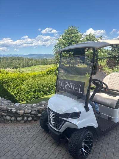 A white golf cart with a black "MARSHAL" sign on its windshield is parked on a paved stone pathway. The cart has plush beige seats and a water bottle is visible on the dashboard. In the background, a meticulously landscaped golf course stretches out with rolling green fairways, bordered by dense trees and distant mountains under a bright blue sky with scattered white clouds. A low stone wall runs along the left side of the pathway, separating it from a lush green hedge. The scene appears to be set in Razlog, Bulgaria, likely at a golf resort given the surroundings. No people are present in the frame.