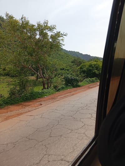 A side-view of a cracked asphalt road from inside a vehicle is presented. The road has numerous visible cracks. To the left of the road, there is a dirt shoulder. Beyond the shoulder, a grassy area transitions into a dense forest covering rolling hills. The sky is clear and pale. The right side of the image is dominated by the interior of the vehicle, showing a dark fabric, possibly a seat or clothing, and the window frame. The location is specified as Rimin Ziem, Nigeria.