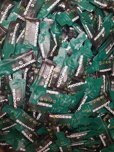 The image shows a large pile of small, individually wrapped green and black packets of what appears to be an energy drink or supplement. The packets are densely packed, obscuring any background details. The text "SUCCESS DAY" is prominently displayed on many of the packets in white lettering. There are also smaller inscriptions such as "ENERGY BOOST," "NATURAL PRODUCT," and "VITAMINS." Some packets have circular logos with symbols like a globe and a stylized plant. The packets appear wet, possibly from condensation or being handled. There are no people visible in the image. The scene is an indoor pile of product packaging, and there is no information that allows for localization within the city of Бұлақты, Kazakhstan.