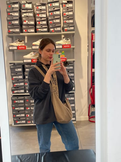 The image shows a woman taking a selfie in what appears to be a retail store in Marcianise, Italy. The reflection suggests she is standing in front of a display of New Balance shoe boxes. There are several shelves filled with these boxes, many with red price tags on them. There are also shoes displayed on the shelves. The woman is wearing jeans and a black top with a light brown cross-body bag. She is holding a smartphone with a green case to take the photo. A red fire suppression system is visible near the ceiling.