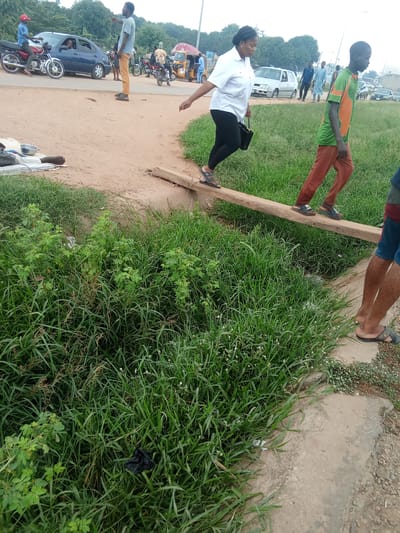 This image depicts a scene in Kagini, Nigeria, likely in a rural or semi-urban area. In the foreground, a wooden plank serves as a makeshift bridge over a ditch filled with lush green grass and some vegetation. Two individuals are walking across this plank. The person on the left, wearing a white shirt and black leggings, is a woman with dark hair pulled back. The person on the right, a younger male, wears a green and orange shirt and brown pants. Their focus is on navigating the unstable crossing.
Further in the background, a dusty road is visible, with several vehicles and people. A blue car is parked on the side of the road, and a person is sitting on a motorcycle in front of it. Another motorcycle, with a rider and passenger, is seen further down the road, alongside a yellow and red structure that appears to be a kiosk or food stall. Several other individuals are walking around this area, and more vehicles are present in the distance. The environment suggests a busy, outdoor setting with elements of both urban and rural life. The overall impression is one of everyday life and movement within the Nigerian town.