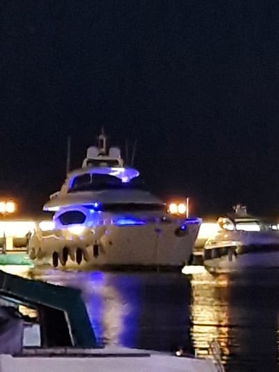 The image depicts a nighttime scene at a marina in Sveti Vlas, Bulgaria. Two yachts are visible, with the primary focus on a larger yacht in the foreground. This yacht is illuminated by blue lights, which are reflected on the dark water. The yacht has a multi-level structure with windows and railings. In the background, a smaller yacht is partially visible, also illuminated by lights. The marina area appears to have a walkway or dock with additional lights. The overall impression is one of a well-lit, upscale waterfront at night.