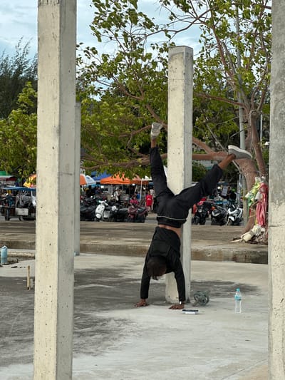 This image shows a person performing a handstand against a concrete beam. The person is wearing black athletic clothing and grey sneakers. They are positioned horizontally, with their hands on the concrete beam and their legs extended upwards. In the background, there are trees, a line of parked motorcycles, and some market stalls. A plastic water bottle is visible on the ground to the right of the person. The scene appears to be outdoors, possibly in a public area or a market in Nathon, Thailand. The sky is visible through the trees, suggesting it is daytime. There is no discernible text in the image.