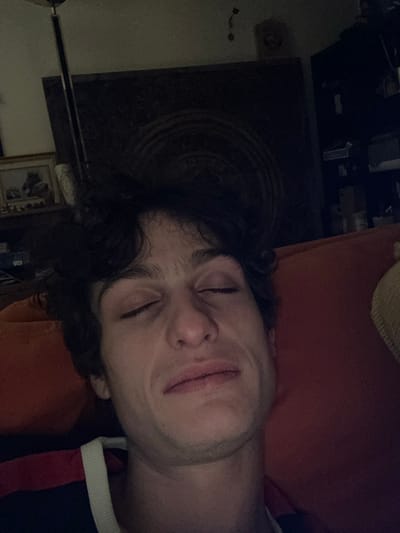 An image taken indoors in Vincennes, France, depicts a young adult male with fair skin and dark, wavy hair. His eyes are closed, and his mouth is slightly open, displaying a neutral to slightly pained or tired expression. He is positioned against a textured orange surface, likely a sofa cushion. A segment of his clothing, a dark garment with a white and red stripe on the collar/shoulder, is visible. The background is dimly lit and out of focus, revealing a floor lamp with a metallic pole, a framed picture on a surface to the left, and a dark shelving unit or furniture piece with indistinct objects to the right. A faint circular pattern is visible on a dark background element behind the subject's head.