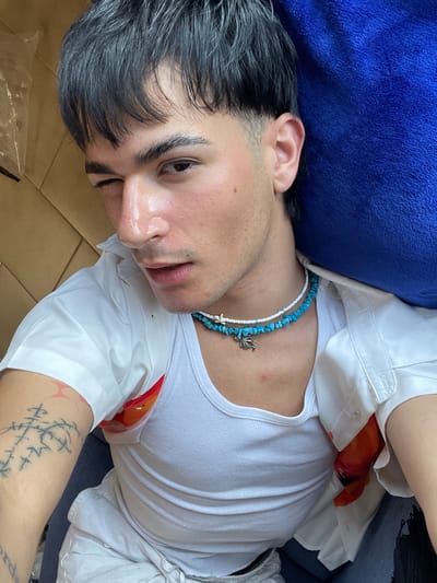 A young person with dark hair is depicted lying down in an indoor setting, likely in Su Masu/Elmas, Italy, bathed in bright natural light, suggesting it is daytime. They are looking towards the camera with a calm and relaxed expression.
The individual is wearing a white ribbed tank top layered under a white short-sleeved button-down shirt. The button-down shirt features an internal orange and red floral or abstract pattern, visible at the armpit and chest area. Around their neck, they wear a casual necklace composed of irregular blue turquoise-like beads interspersed with small white beads, featuring two silver marine-themed charms – one appearing to be a starfish and the other possibly an octopus or crab.
Their dark hair appears thick and slightly styled or damp. Subtle stubble is visible on their chin and jawline. A distinctive black ink tattoo with runic or abstract linear symbols is prominently displayed on their left forearm, accompanied by some red marks on the skin.
The person is resting their head on a dark blue plush pillow or fabric object, while their body lies on a light brown surface, possibly cardboard or a wooden floor. The overall scene conveys a sense of quiet relaxation. There is no visible text in the image.