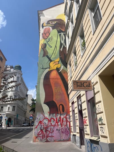 The image shows a building in Vienna, Austria, located at latitude 48.195, longitude 16.346. The building has a large mural painted on the side, depicting an eagle holding a cello. The mural is in a stylized, modern art style. The mural is painted on a wall with a light yellow paint, with a light orange painted background. The cello is a vibrant red, with the rest of the mural painted in shades of green, yellow, and brown. The building next to the mural is a light cream color, with a sign reading "FatCat Tattoo Shop." The tattoo shop is a small store with a glass storefront. The storefront has a sign that is also spray-painted, with red letters on the background. The image is taken from the street, looking towards the building. The ground is covered in pavement, and there are some cars parked on the street. The sky is a bright blue with a few white clouds. There are no visible people in the scene.