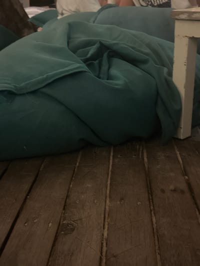 A turquoise comforter, partially draped, sits beside a weathered wooden plank surface. The comforter features several folds and curves. A section of white wooden furniture is also visible near the comforter. The plank surface exhibits natural variations in color and texture, with prominent seams between the planks.