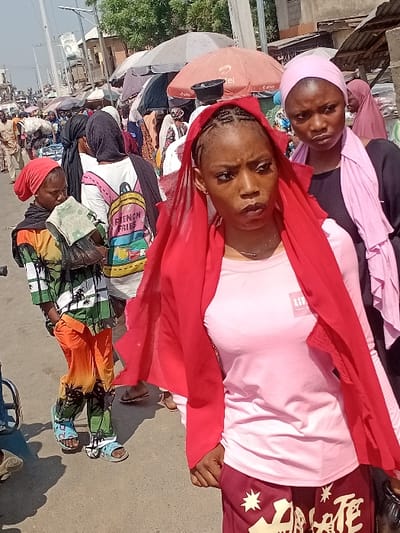 Several people are gathered on a sunny street. Two women take center stage, one in a pink top with a red scarf, the other behind her with a light purple head covering. Market umbrellas dot the background. The atmosphere appears bustling, possibly a market day, with people dressed in a variety of colorful clothing. The image is slightly tilted, offering a candid snapshot of everyday life in an outdoor setting.