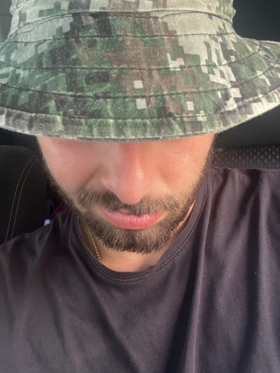 A close-up, chest-up photograph shows a person wearing a camouflage bucket hat and a dark t-shirt. The hat obscures the upper half of their face, with only the lower nose, mouth, and chin visible, covered in stubble. A gold chain is visible around the person's neck. The background is dark and appears to be the interior of a vehicle, with a textured headrest visible to the right. The overall lighting suggests an outdoor setting, possibly during the day. No specific landmarks or details indicate the location in Stepantsminda, Georgia.