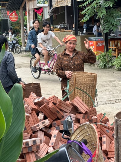 In Mai Chau, Vietnam, a woman wearing a brown patterned shirt and a red and yellow headscarf stands in the foreground, holding a woven basket and gesturing with her right hand. To her left, a pile of red bricks is visible, partially obscured by large green leaves. Behind her, two young men are riding a pink bicycle. The man in front, wearing a denim jacket, smiles, while the man behind him, wearing a white t-shirt and glasses, also smiles. In the background, buildings with wooden structures and signage are present. A motorcycle is parked to the left, and a bright orange cooler with the "Merino" logo is visible. Lush green plants and trees contribute to the outdoor environment. The scene appears to capture a moment of daily life in a Vietnamese town.