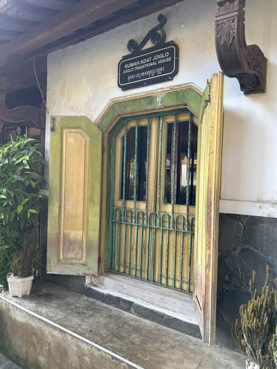This media file shows an exterior view of a traditional Joglo house. The primary subject is a large, open window with green and yellow painted wooden frames and protective metal bars. The window is flanked by decorative wooden architectural elements. A black sign with white text is affixed to the wall above and to the left of the window. The sign reads: "RUMAH ADAT JOGLO TRADITIONAL HOUSE". There appears to be Javanese script below the English text.

The scene is set outdoors, with a stone pathway or ground visible in the foreground and to the right. Green foliage from plants in pots is present at the bottom of the frame, and dried, spiky branches are visible at the top right, possibly part of a decorative planting or natural growth. The wall of the house has peeling paint, suggesting age and exposure to the elements. The lighting suggests it is daytime, but the specific time of day is unclear. The overall impression is of an old, culturally significant building.