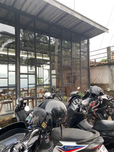 The image shows a view of a building with a large glass facade, in Kecamatan Piyungan, Indonesia. The building is constructed with a corrugated metal roof. The windows are framed in black metal and divided into numerous panes. Behind the building, there is a view of a forested area. The building is empty, but four motorcycles are parked in front of it. The motorcycles are parked along a walkway, and all are facing the street. The motorcycles are black and red, and they are all equipped with helmets. One of the helmets is black and has a sticker on it that reads "KCP 0328". There is a sign on the motorcycle that reads "BEAT" in red. The sign is located on the side of the motorcycle and is written in white. The motorcycle is facing the viewer. The building is located at -7.855 latitude and 110.473 longitude.