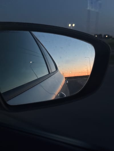 A side mirror view shows a car on the road at dusk or dawn. Street lights are reflected in the side mirror. Based on image characteristics, it is not possible to determine the location in Mamadysh, Russia.
