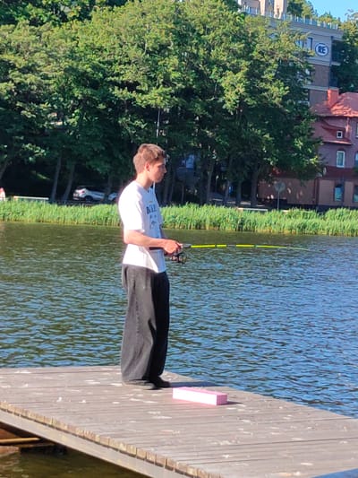 A young person stands on a wooden pier, fishing in a body of water. They are wearing a white t-shirt with a graphic print and loose black pants. The person holds a fishing rod with a yellow and green line extending into the water. The pier is made of weathered wooden planks. In the background, there is a line of tall green trees along the water's edge. Beyond the trees, buildings are visible, including a modern building with a blue circular logo containing the letters "RE". The scene suggests a warm, sunny day, likely in the late morning or early afternoon, given the bright sunlight and clear sky. The water is rippled, indicating a gentle breeze. A bright pink rectangular object rests on the pier near the foreground. The setting appears to be a recreational area in Svetlogorsk, Russia, possibly a lake or a wide river. The overall mood is peaceful and focused on the activity of fishing.