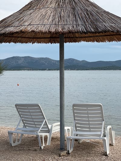 This image depicts a tranquil beach scene in Betina, Croatia. In the immediate foreground, a large, circular thatched straw umbrella with a central grey metal pole provides shade over a gravel and pebble beach. Positioned under the umbrella and facing the water are two empty white plastic lounge chairs or recliners, one on the left and one on the right, both slightly angled towards the center. Beyond the beach, a calm body of water, likely the Adriatic Sea or a bay, extends towards the horizon. An orange spherical buoy floats in the water to the left of the central pole, and a horizontal line of smaller white buoys or floats stretches across the mid-distance of the water. In the background, a series of dark, undulating hills or mountains covered with sparse vegetation define the distant coastline. The sky above is overcast or hazy with a light blue-grey tone. No individuals are present in the scene.