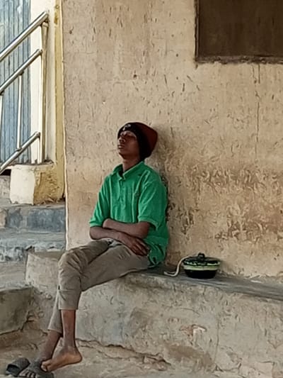 In Arawa, Nigeria, a young male with dark skin, wearing a maroon beanie and a green polo shirt, sits with his arms crossed on a concrete ledge. His eyes are closed, suggesting he is resting or contemplating. He is wearing light brown pants and dark sandals. Beside him on the ledge sits a small black container with a white string. The background consists of a textured, light-colored wall and some steps with a metal railing.