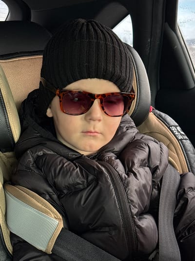 The scene is situated within a vehicle, consistent with the specified location of Bol'shaya Sosnova, Russia. A young child, appearing to be male, is seated in a forward-facing child safety seat. The child wears a black ribbed knit beanie and tortoiseshell-patterned sunglasses with reddish-purple tinted lenses, obscuring the eyes. A black puffy, quilted jacket with a visible zipper is worn. The child's expression is neutral and serious, with lips closed. The child safety seat features a two-tone design, with beige/tan fabric on the headrest and side bolsters, and black fabric elsewhere. A black seatbelt with a beige/light grey padded cover is fastened across the child's chest. The vehicle's interior appears dark, with black upholstery and a partial view of a window showing a blurred exterior on the upper right.