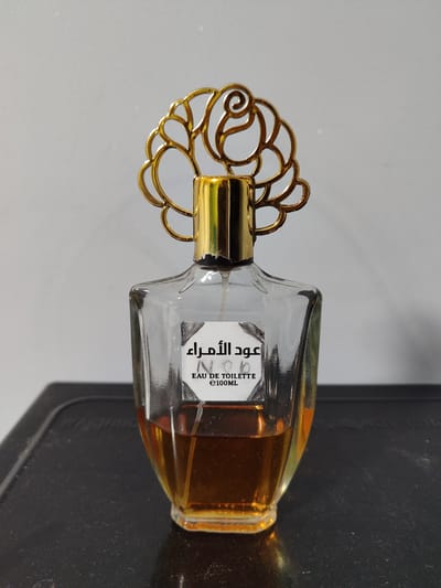 A clear glass perfume bottle with a gold ornate cap sits on a black surface. The bottle contains amber-colored liquid. A white label on the front of the bottle has text in Arabic and English, reading "عود الأمراء EAU DE TOILETTE e100ML". The background is a plain grey wall. This image was taken in the city of Sudak, Ukraine.