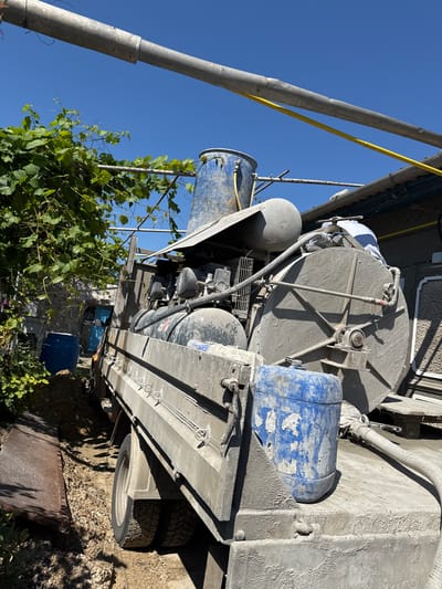 This image shows a concrete mixer truck parked outdoors, likely at a construction site in Ștefan Vodă, Moldova. The truck is the primary subject, with its large mixing drum and associated machinery visible. Several blue barrels, one of which is heavily weathered, are also present. Green foliage, appearing to be grapevines, is growing in the foreground, suggesting a rural or semi-rural setting. The scene is bathed in bright sunlight, indicating it is daytime. The sky is a clear, vibrant blue, suggesting fair weather. Shadows are cast by the truck and the foliage, further emphasizing the sunny conditions. The truck itself appears to be in use or recently used, as there are visible signs of concrete dust and residue on its surfaces. No people are visible in the frame. The overall impression is one of an active construction environment on a sunny day.