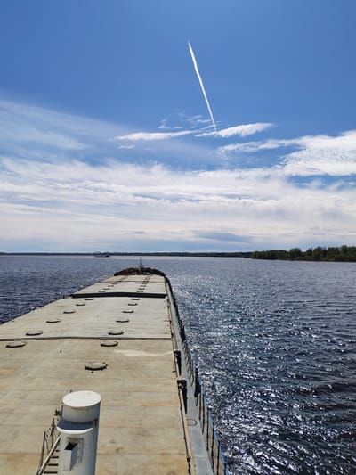 The image shows a view from a boat or dock, looking out over a body of water. A flat, concrete surface with hatch covers stretches out into the water. To the right and left of this surface is the water, sparkling in the sunlight. In the distance is a shoreline with trees and possibly a bridge. The sky is blue with scattered clouds and a contrail from an airplane. The location is identified as Glebovo, Russia.