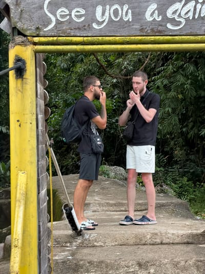 Two young adult males are standing on a set of concrete stairs. The male on the left is wearing a black t-shirt, black shorts, a black backpack, and black and white patterned flip-flops. He has sunglasses perched on his head and is holding his hand to his mouth. The male on the right is wearing a black polo shirt, white shorts, and dark blue Crocs. He has a small black crossbody bag slung over his shoulder and is holding his hands up near his chin. Both individuals have short hair. They are standing under a wooden sign that reads "See you again" in white lettering. A yellow metal structure with a chain hanging from it is to the left of the frame. The background consists of dense green foliage, suggesting an outdoor, possibly tropical, environment. The scene appears to be in Khanh Vinh, Vietnam, based on the information provided.