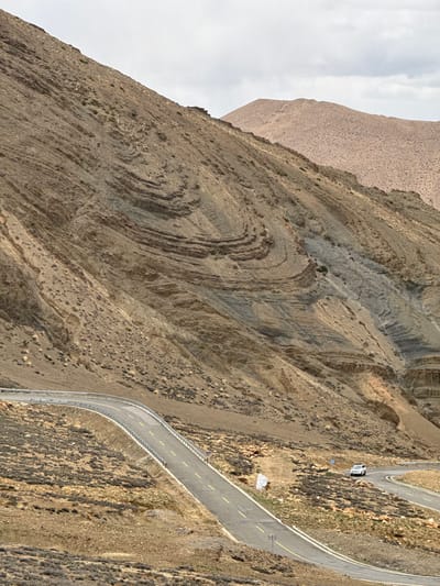 The image shows a winding asphalt road traversing a steep, arid mountainside. The road has yellow lines and guardrails along its edge. A single, light-colored van is parked on a flat area adjacent to the road. The mountain exhibits distinct geological features, including folded rock layers and erosion patterns. The surrounding environment appears barren with sparse vegetation and gravel. The town of Zagor is not visible. It's located in China.